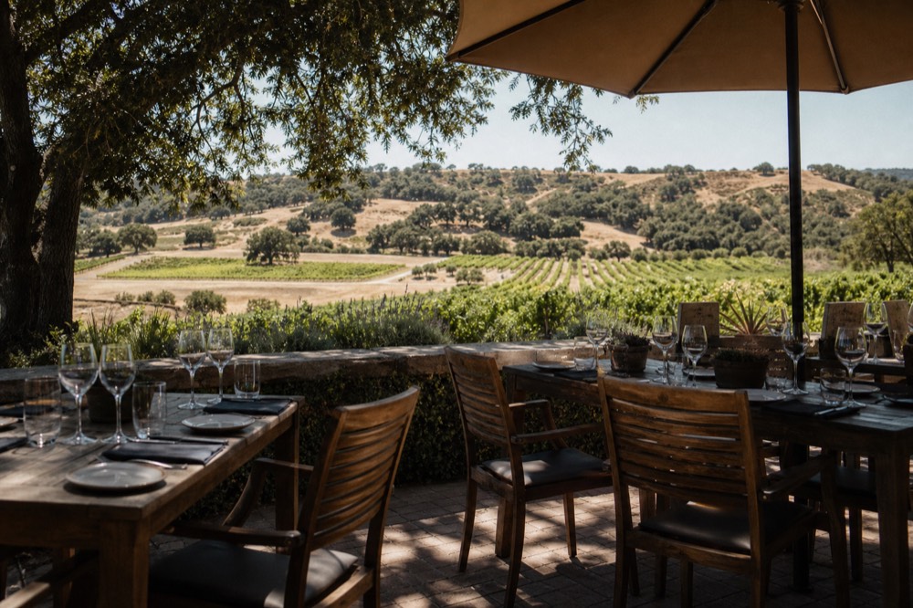 A seated estate tasting terrace with reserved place settings and wine glasses overlooking vineyard rows and distant oak hills, no guests yet, warm afternoon light, no text, no logos, no people