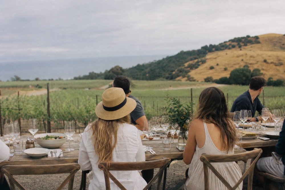 A group seated at a long vineyard table sharing a lunch and wine glasses with vines and golden hills behind, everyone relaxed in afternoon light, no text, no logos, no people