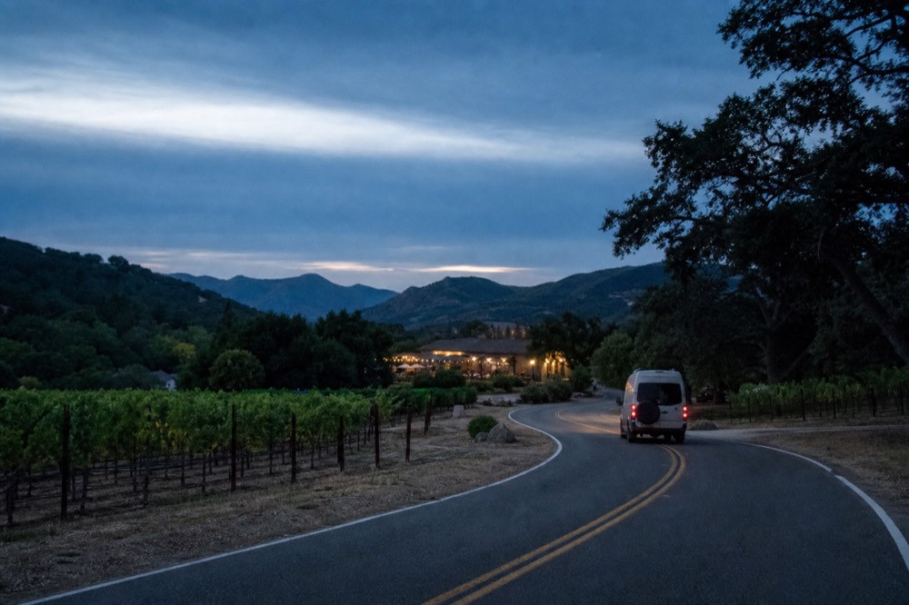 A small touring van on a two-lane valley road winding between vineyard blocks and oak trees under a wide sky, no text, no logos, no people