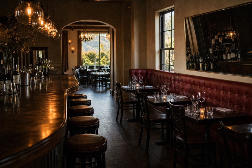 A dim warm-lit steakhouse interior with a long polished bar, red banquettes and wine glasses set on dark wood tables, no text, no logos, no people