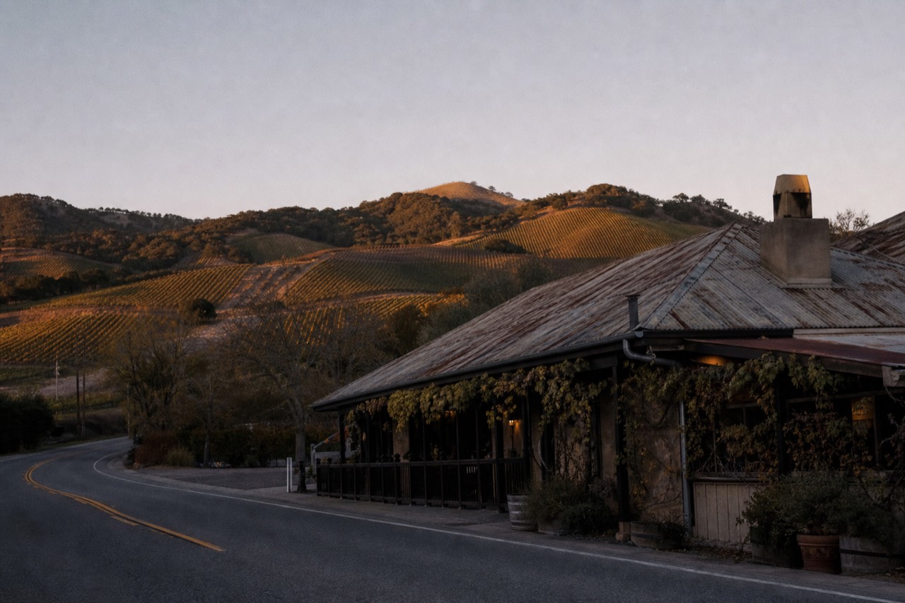 A weathered roadside restaurant and bar building at dusk beside a two-lane highway in the Santa Ynez Valley with vineyard hills behind, no text, no logos, no people