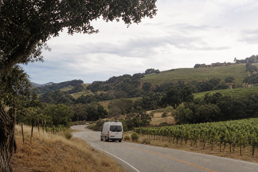 A small touring van on a two-lane valley road winding between vineyard blocks and oak trees under a wide afternoon sky, no text, no logos, no people