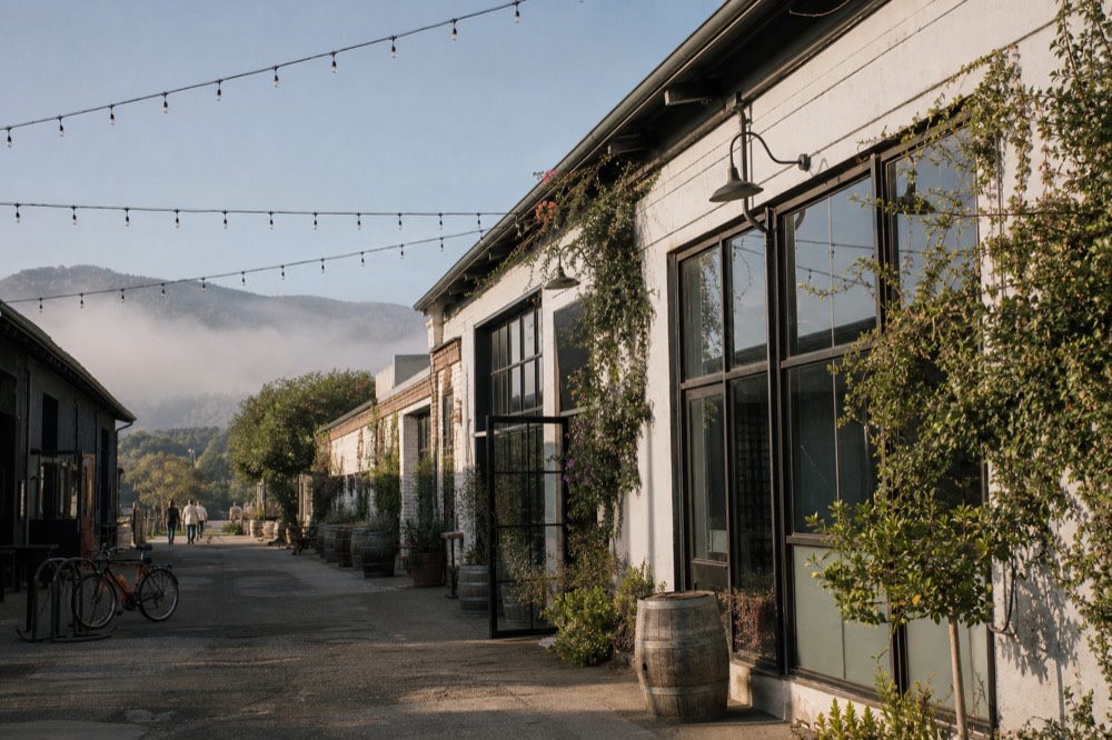 A row of converted-warehouse tasting rooms along a Funk Zone lane with string lights overhead and a couple of parked bikes, no text, no logos, no people
