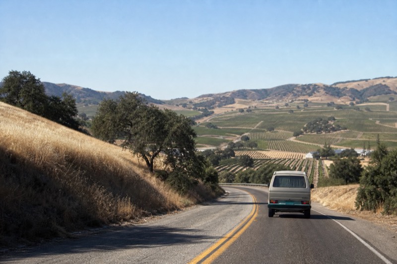 A small touring van on a two-lane road climbing between golden hills toward the Santa Ynez Valley vineyards under a clear California sky