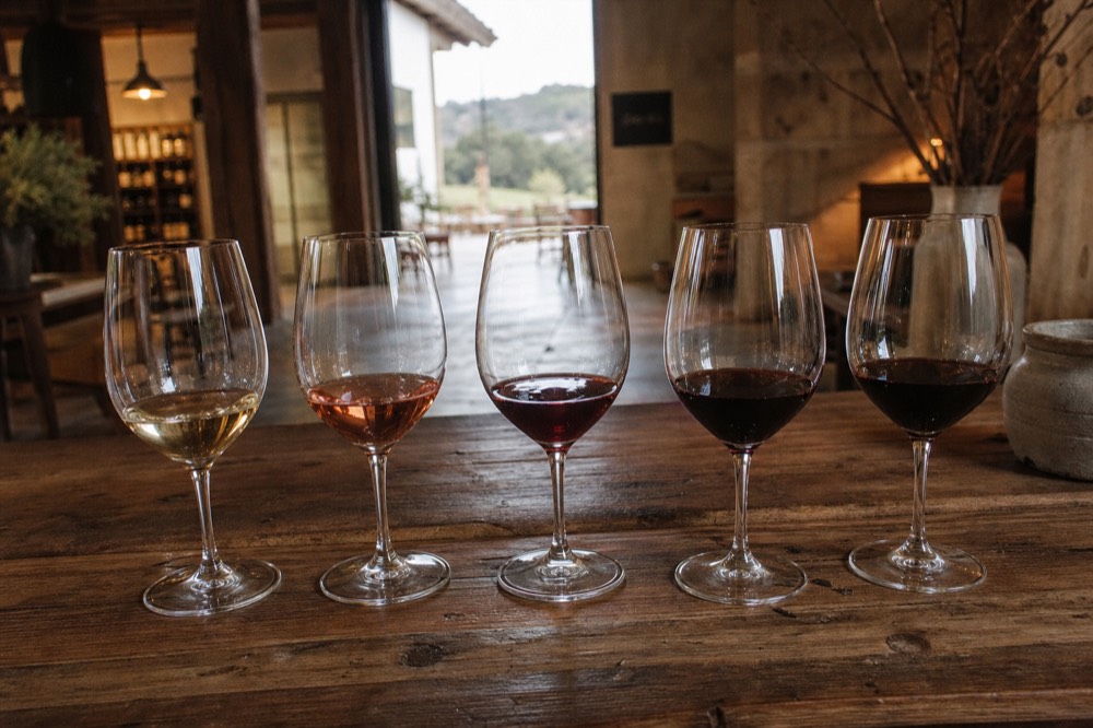A tasting flight of five wine glasses arranged on a rustic wooden counter inside a warm-lit village tasting room, no text, no logos, no people