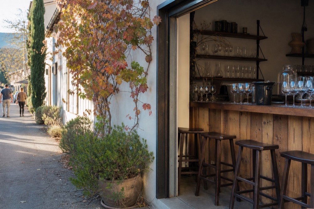 An open tasting-room doorway framing a wooden bar and shelves of glassware on a shaded village street, no text, no logos, no people