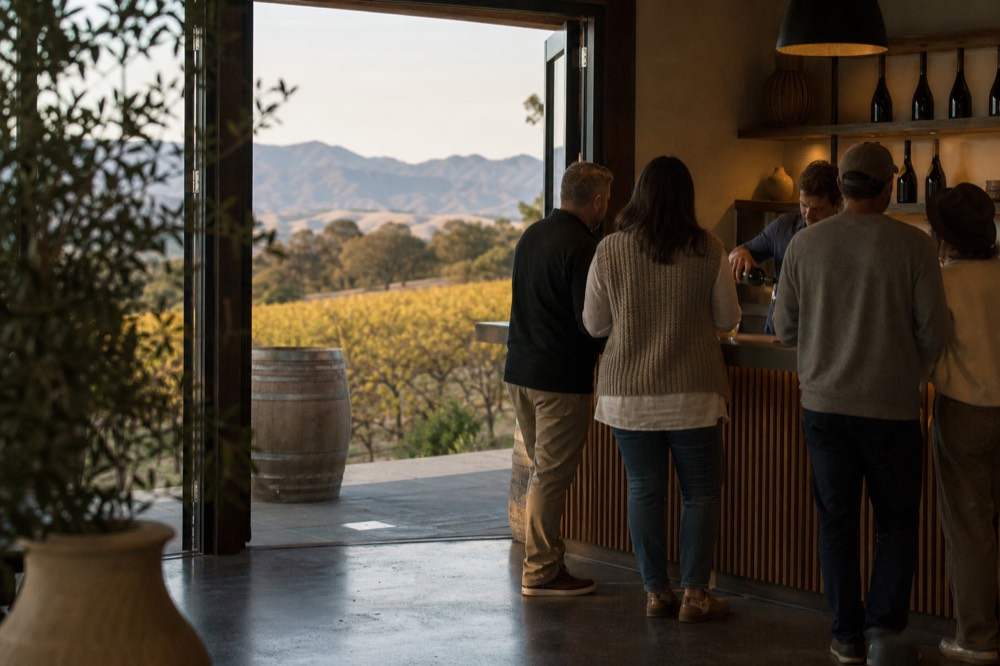 A small group standing at a tasting-room counter while a host pours, warm interior light and wine bottles on shelves behind, no text, no logos, no people