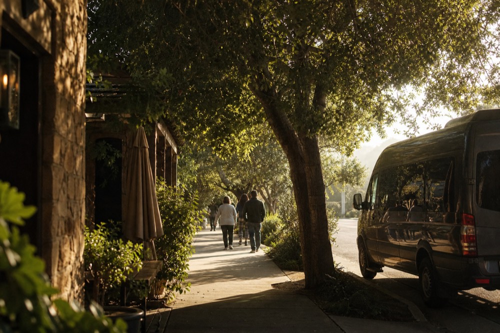 People walking a shaded village sidewalk between tasting-room storefronts with a parked touring van at the curb in warm light, no text, no logos, no people