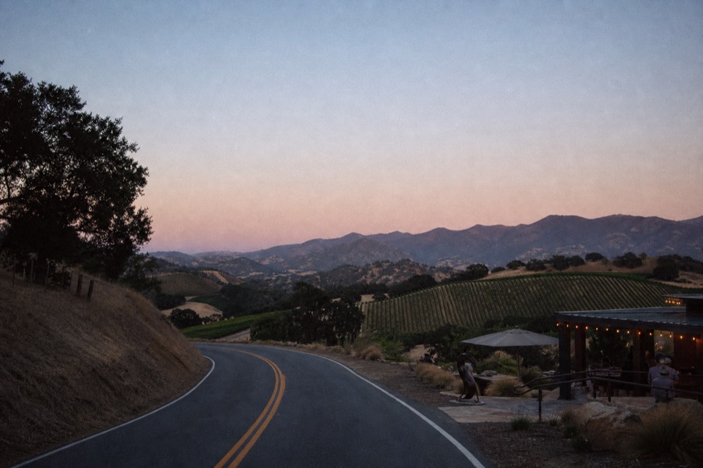 A winding two-lane road climbing over a mountain pass with oak-studded golden hills and vineyards visible in the valley below, no text, no logos, no people
