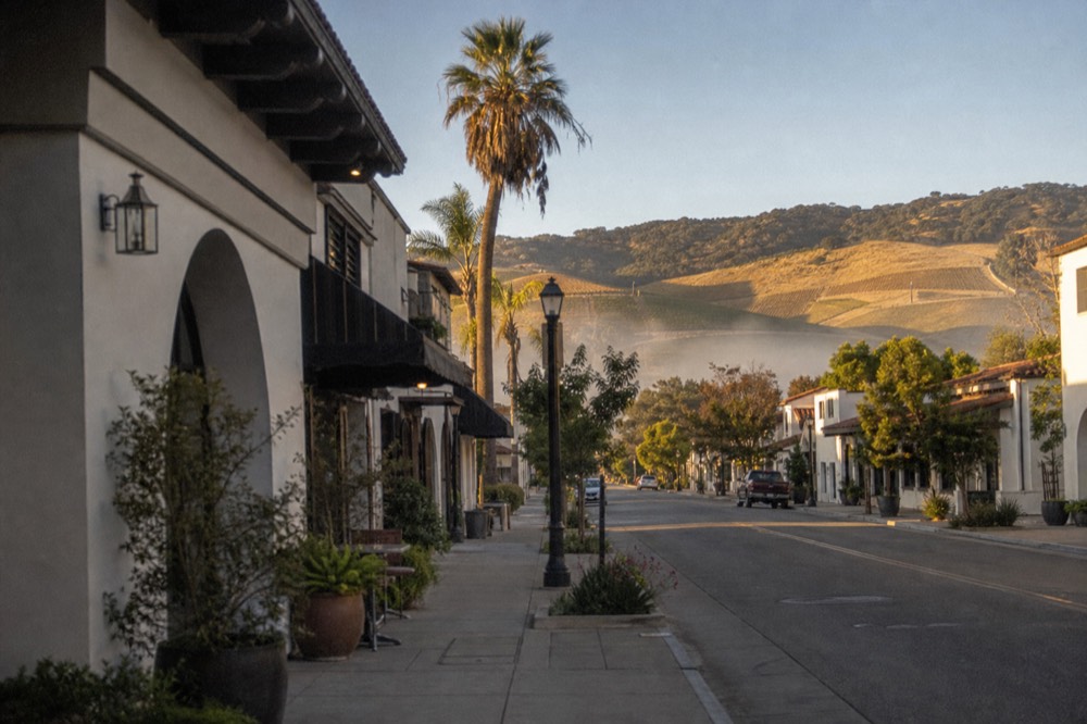 A split view feel of a coastal city street of tasting rooms giving way in the distance to golden inland vineyard hills under a wide California sky, no text, no logos, no people