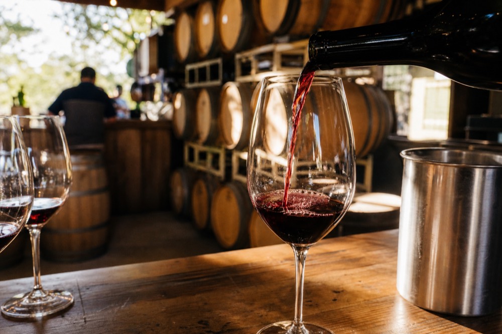 A wine pour caught mid-stream into a glass at a rustic cellar-door counter with barrels blurred behind, warm light, no text, no logos, no people