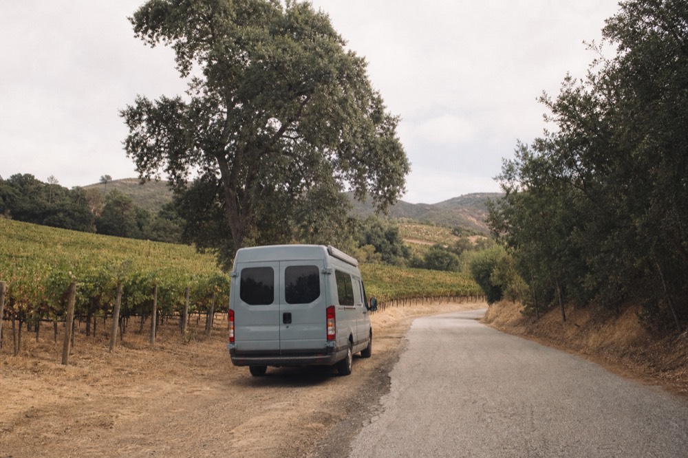 A small touring van parked on a quiet Santa Ynez Valley road between vineyard blocks and oak trees in afternoon light, no text, no logos, no people
